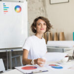 Beautiful smiling girl with short curly hair in wireless earphones and white T-shirt happily looking in camera working on laptop with cup of coffe near at work in modern cozy office